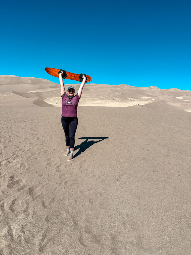A girl in a maroon t-shirt holding a sandboard over her head while camping at the Great Sand Dunes