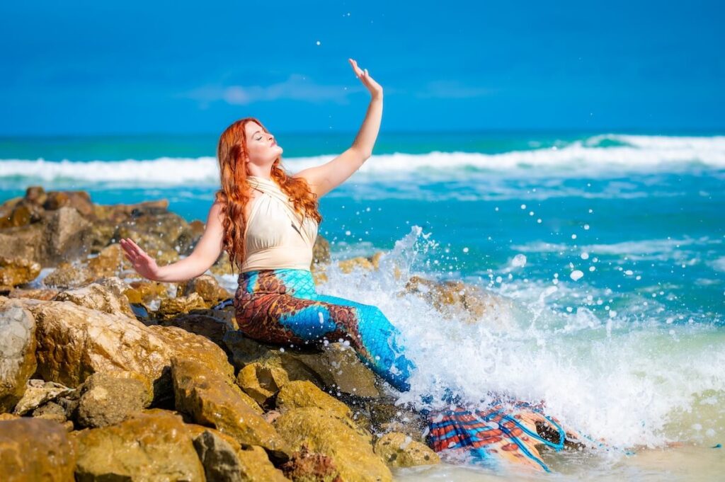 A mermaid with a blue and orange tail on a rock with waves splashing over her