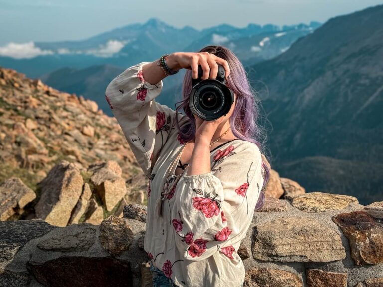 a photographer with purple hair holding up her camera with a mountain landscape behind her.