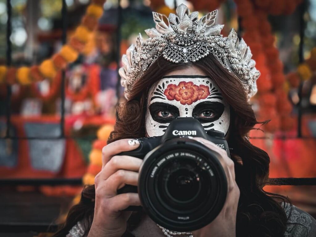 A red haired girl with a crown in day of the dead, scary carina makeup, holding up a camera in front of her face in Sante Fe New Mexico