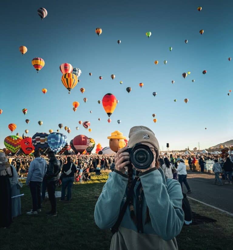 A girl with a beanie holding a camera towards the lens with hundreds of balloons behind her at the Albuquerque Balloon Festival