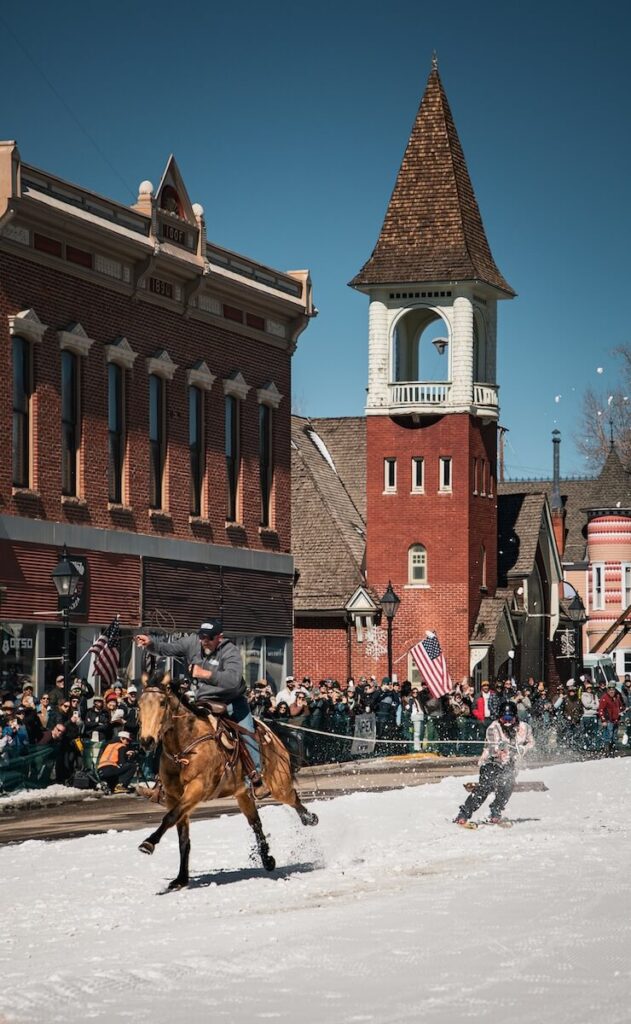 A brown horse and cowboy pulling a skier for skijorning through the town of Leadville, Colorado