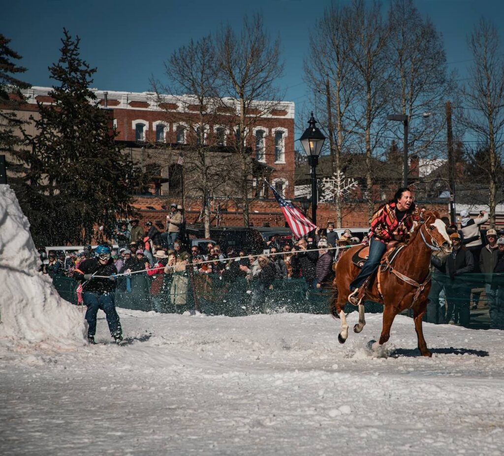 A brown horse pulling a skier off of a jump for skijoring in Leadville, Colorado with a crowd watching