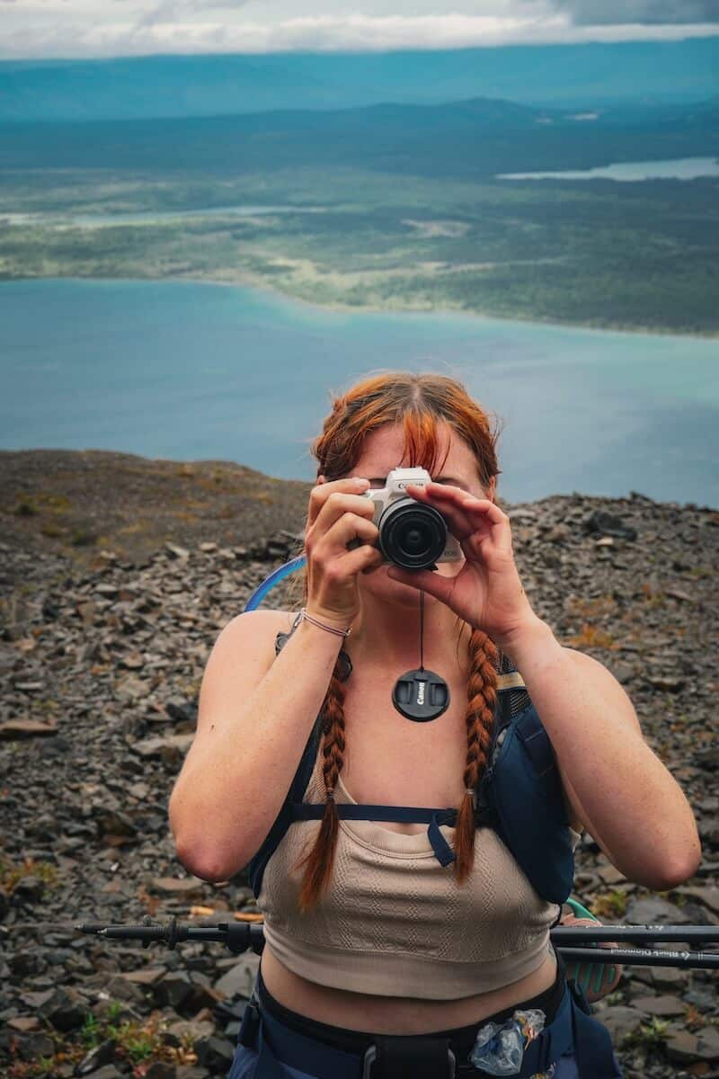 A red-haired girl in a pink tank top hiking, holding a white camera up to her face, with Kathleen Lake behind her while hiking King's Throne in Kluane National Park in Canada